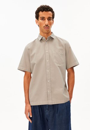 Mann trägt ein beige kurzärmeliges Hemd mit Brusttasche und dunkelblaue Jeans vor einem schlichten weißen Hintergrund.