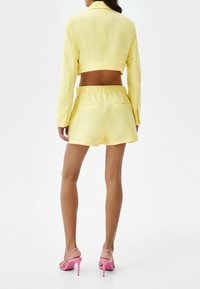 Woman wearing yellow cropped blazer and matching elastic waist shorts with pink high-heeled sandals, shown from behind on white background.