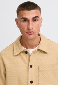 Beige ribbed shirt jacket with a spread collar, black buttons, and a front pocket, made from textured fabric.