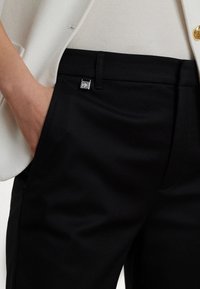 Svarta byxor med en slät yta har en silverfärgad logoknapp och fickor, i kombination med en ljusfärgad, strukturerad blazer.