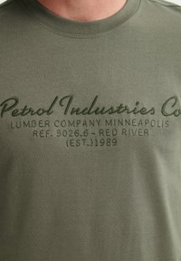 Tricou de culoare verde măsliniu, fabricat din bumbac moale, cu text brodat în verde mai închis. Detalii notabile includ brandingul și anul de înființare.