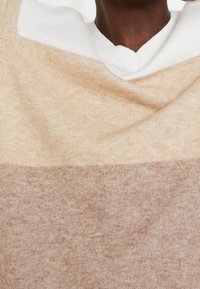 Maglione lavorato a maglia con un design a blocchi di colore orizzontali in crema e marrone chiaro. Tessuto morbido, scollo a V con bordo a costine e dettagli hardware minimali.