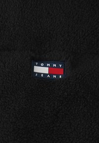 Svart fleece-tyg med en texturerad yta. Har en rektangulär logotyp med blå, röd och vit färg med texten "TOMMY JEANS."