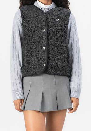 Personne portant un pull gris à torsades, un gilet noir en peau lainée avec des boutons, et une jupe plissée grise, debout devant un fond blanc uni.