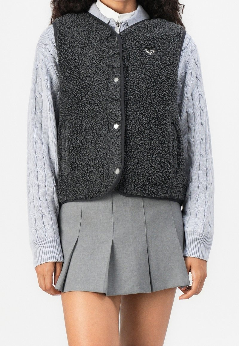 Personne portant un pull gris à torsades, un gilet noir en peau lainée avec des boutons, et une jupe plissée grise, debout devant un fond blanc uni.