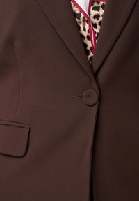 Bruine blazer met een zachte textuur, voorzien van een ronde knop, notch revers en een enkel borstzakje. Voering zichtbaar eronder.