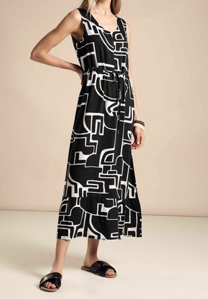 Frau trägt ärmelloses Midi-Kleid mit schwarz-weißem geometrischem Muster, Kordelzug in der Taille und schwarzen Sandalen zum Hineinschlüpfen.
