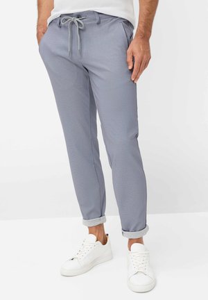 Mand iført lysegrå snørede bukser med manchetter ved anklerne og hvide sneakers, stående mod en ensfarvet baggrund.