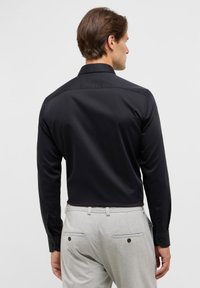 Maglietta nera a maniche lunghe con una texture liscia, dotata di un colletto classico e polsini con sopra il bottone, indossata con pantaloni a quadri grigio chiaro.