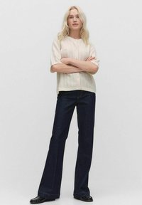 Crème kleurige cardigan met korte mouwen, kabelbrei-textuur en parelknopen, gecombineerd met donkerblauwe wijde jeans en zwarte schoenen.