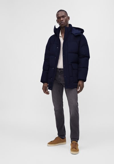 Homme portant une veste matelassée bleu foncé, une chemise beige clair, un jean gris et des baskets marron, debout devant un fond uni gris clair.