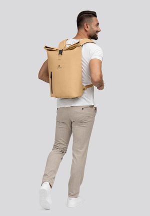 Mochila beige impermeable con tapa enrollable, con un logo, correas ajustables y un bolsillo con cremallera lateral, llevada por un hombre con atuendo casual.