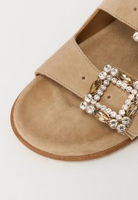 Beige mockasinsandaal med en bred rem och en smyckad spänne med klara och ljusbruna stenar, med en platt sula och mjuk textur.