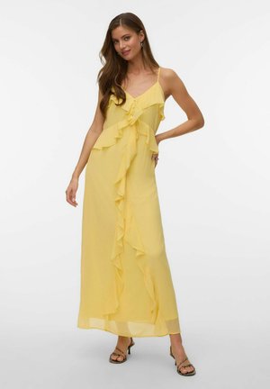 Vrouw in een gele mouwloze maxi-jurk met ruches en spaghettibandjes, draagt sandalen met hakken tegen een effen witte achtergrond.