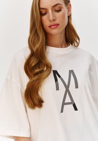 Bia�ła, oversize t-shirt z czarnymi graficznymi literami "MA" w różnorodnych wzorach. Miękki materiał bawełniany, okrągły dekolt i krótkie rękawy.