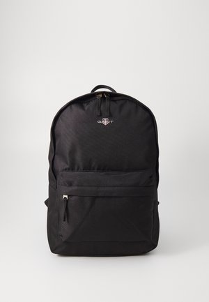Crni ruksak od tkanine za koji je karakterističan zaobljen oblik, s prednjim džepom na zip i ručkom na vrhu. Sadrži logotip brenda sprijeda.