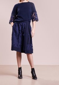 Laivastonsininen paita, jossa on kukkakuvioiset pitsihihat, yhdistettynä laivastonsiniseen hameeseen, jossa on teksturoituja kukkakuvioita ja sidottava vyötärö. Mustat nilkkurit.