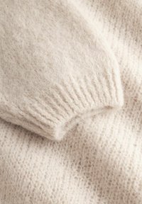 Pehme kergekreemjas kootud kangas, mis näitab lähedalt soonikuga manseti detaili udusel kampsuni varrukal.