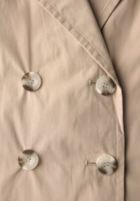Beige op maat gemaakte jas met een dubbelzijdig ontwerp, voorzien van grote, doorzichtige knopen en een gladde stof met een lichte glans.
