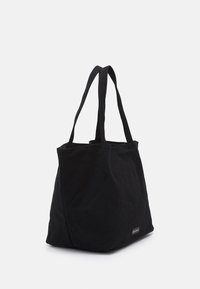 Černá plátěná taška s dvěma dlouhými uchy, prostorným designem a malou značkovou etiketou na boku, která má měkkou texturu.