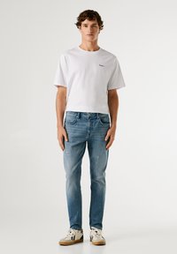 Camiseta blanca de manga corta con logo pequeño; jeans ajustados de color azul claro con ligero desgaste; zapatillas beige con acentos negros y coloridos.