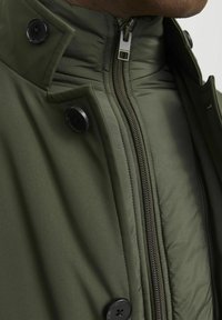 Giacca imbottita verde oliva con colletto alto, dotata di una chiusura frontale con cerniera e due bottoni neri al collo per la chiusura.