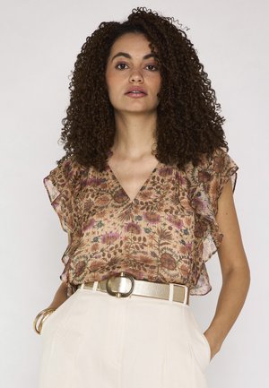 Donna con capelli ricci che indossa una camicetta di pizzo floreale, pantaloni color crema a vita alta, cintura dorata e braccialetto dorato, in piedi con le mani nelle tasche.