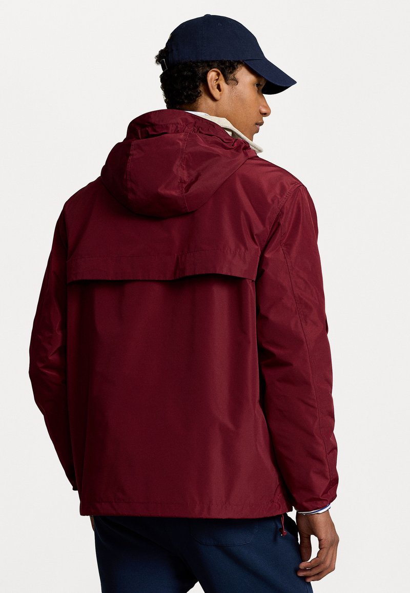 Bordeauxrote, wasserdichte Jacke mit Kapuze, ausgestattet mit einer Rückenpasse und seitlichen Reißverschlusstaschen. Hergestellt aus glattem, leichtem Material.