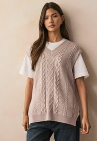 Pulover tricotat fără mâneci, de culoare roz deschis, cu decolteu în V, având modele texturate și detalii de tricot cu fire răsucite, suprapus peste un tricou alb.