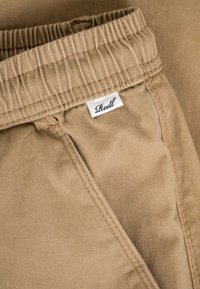 Nahaufnahme des beigen elastischen Bunds und der Fronttasche einer Hose mit einem weißen Stoffetikett der Marke "Reell".