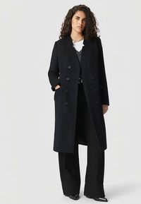 Lange zwarte wollen jas met dubbele rij knopen, zijzakken en een rechte silhouet, gedragen over een gelaagde zwarte outfit.