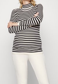 Maglione a collo alto a strisce bianche e nere con texture a coste, caratterizzato da maniche lunghe e un design aderente, abbinato a pantaloni bianchi a coste.