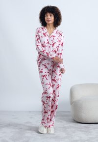 Žena stojí v bielych pyžamách s červenými vzormi mašlí a bielych papučiach, vedľa bieleho čalúneného kresla.