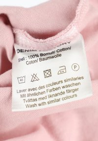 Label on light pink fabric with care instructions. Text in multiple languages. Material is 100% cotton. Care symbols are clearly visible.
