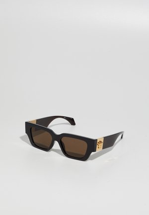 Lunettes de soleil noires rectangulaires avec des verres brun foncé et des détails dorés sur les branches, posées sur une surface gris clair.