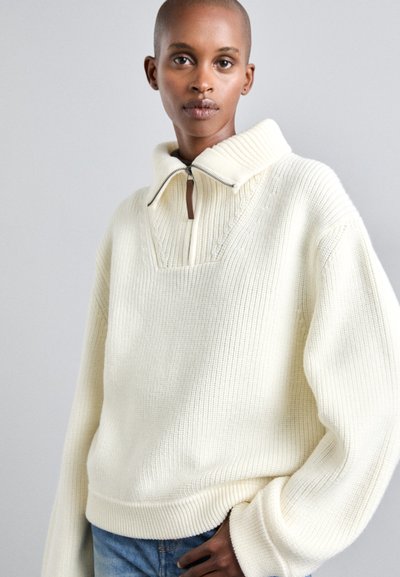 Pulover oversized de culoare crem, tricotat, cu guler înalt, fermoar pe jumătate și textură cu rigle, asortat cu blugi din denim albastru.