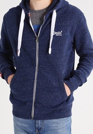 Mann trägt marineblaue Kapuzenjacke mit Reißverschluss und weißen Kordeln, Hände in den vorderen Taschen und graues Shirt darunter.
