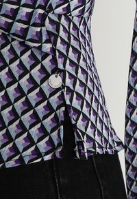 Tryckt topp i lättviktigt tåligt tyg med ett geometriskt mönster av lila, blå och svarta former. Inkluderar en metall-logotagg.