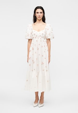 Kvinde med langt mørkt hår iført en hvid blomsterprintet midi-kjole med pufærmer og hvide hæle, stående mod en ensfarvet baggrund.