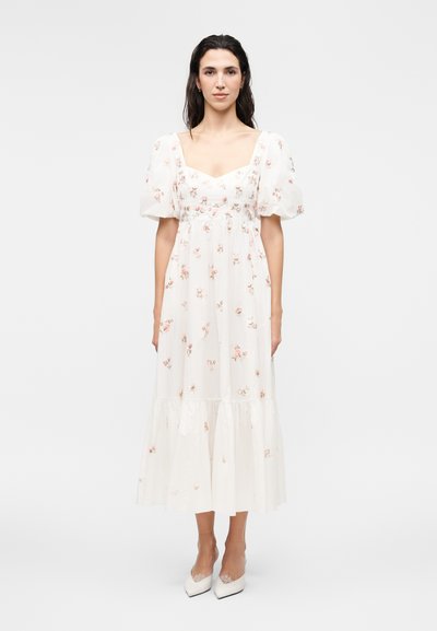 Woman with long dark hair wearing a white floral midi dress with puff sleeves and white heels, standing against a plain background.