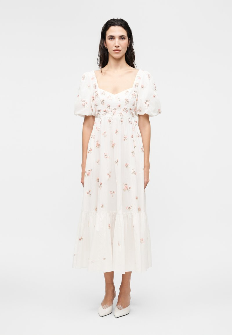 Γυναίκα με μακριά σκουρόχρωμα μαλλιά, που φοράει ένα λευκό φόρεμα μέσης μήκους με λουλούδια, φουσκωτά μανίκια και λευκές γόβες, στέκεται μπροστά σε απλό φόντο.