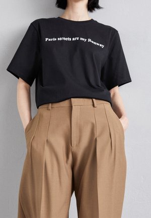 Persona con una camiseta negra con el texto "Las calles de París son mi pasarela" y pantalones marrones de cintura alta y plisados, con las manos en los bolsillos.