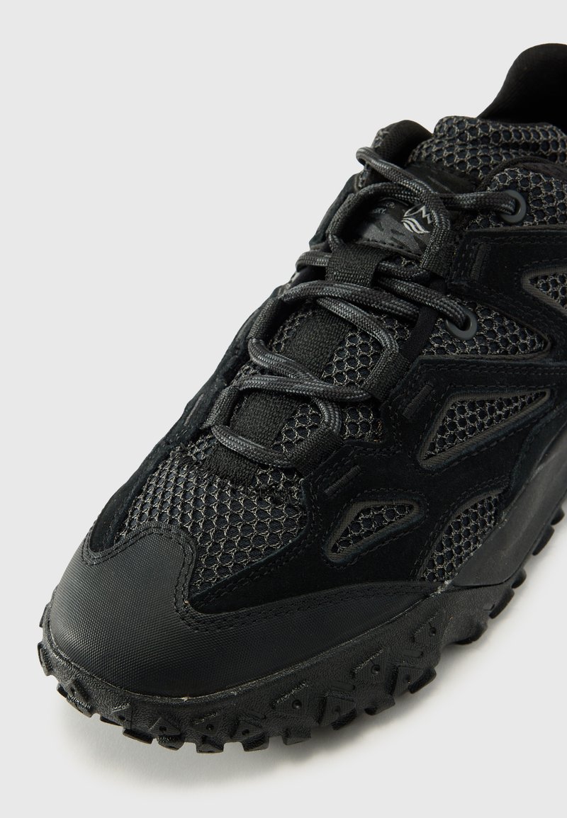 Zapatilla deportiva negra con parte superior de malla texturizada, suela de goma y cierre de cordones. Presenta una puntera redondeada y un patrón de relieve distintivo.