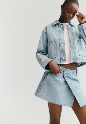 Giovane donna che indossa una giacca di jeans azzurro chiaro e una gonna asimmetrica abbinata, che posa con una mano in tasca e l'altra che si tocca la fronte.