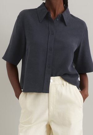 Persoon draagt een donkergrijs, kort mouw, overhemd met knopen, lichtjes ingestopt in een hoog getailleerde, gebroken witte broek met handen in de zakken.