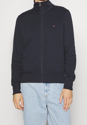 Mężczyzna w granatowej kurtce zapinanej na suwak z wysokim kołnierzem i jasnoniebieskich dżinsach, stojący na jednolitym tle.