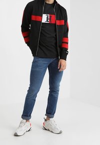 Chaqueta negra con cremallera y rayas rojas, camiseta negra con gráfico blanco, jeans ajustados azules y zapatillas blancas con acentos grises y naranjas.