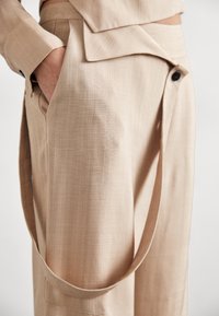 Beige Schnitthose aus strukturiertem Stoff mit seitlichem Taschen und einem langen Tragegurt, der von der Taille herabläuft.