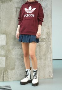 Felpa bordeaux con logo Adidas bianco, mini gonna in denim plissettata e stivali da combattimento bianchi con lacci e dettagli neri, con suole spesse.