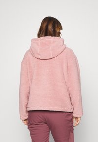 Růžová fleecová bunda s kapucí. Měkký materiál, volný střih a viditelné švy podél okrajů a kapuce.